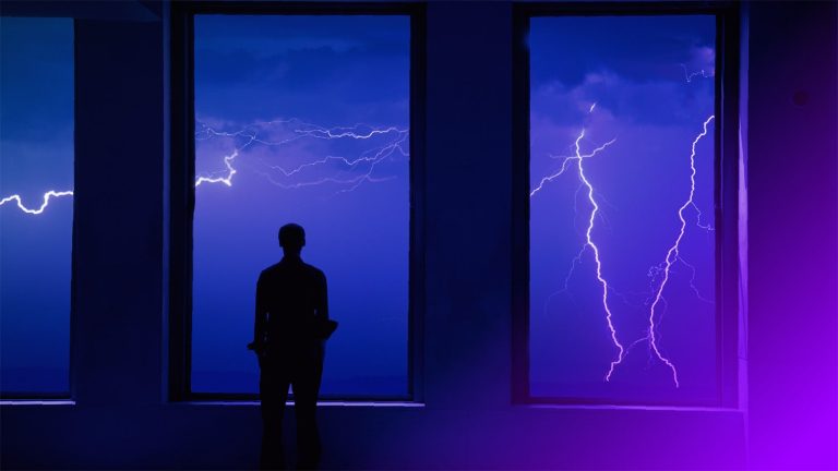 A man stares out a window at a lightning storm.